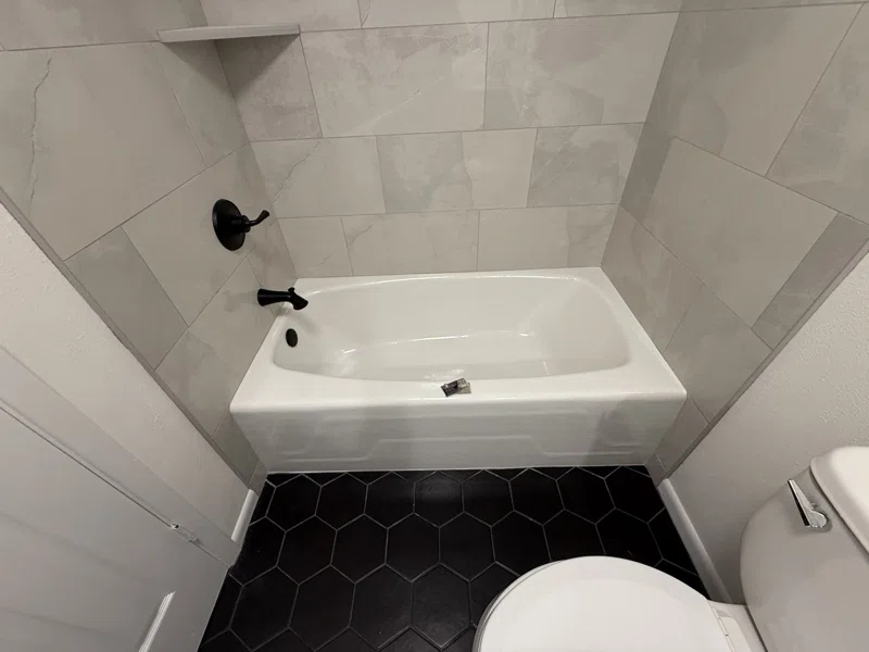White bathtub with black fixtures, a tiled surround and dark hexagonal floor tile.